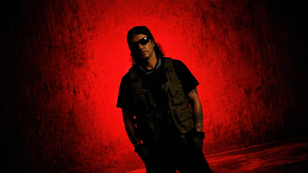 Man wearing sunglasses, a beanie, and a tactical vest against a textured red background