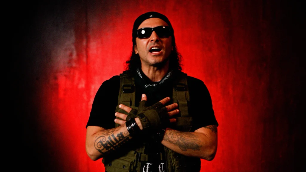 Rapper Killa Hakan wearing sunglasses and a tactical vest with hands crossed over his chest against a red background