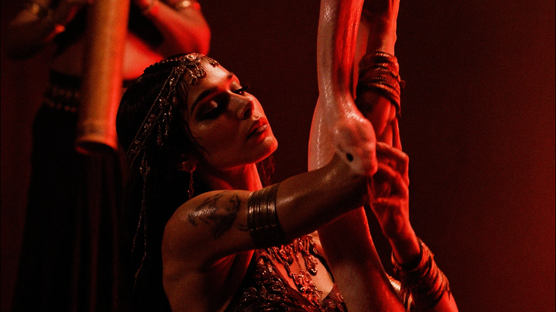 A woman in ornate jewelry holds a large albino snake under dramatic red lighting