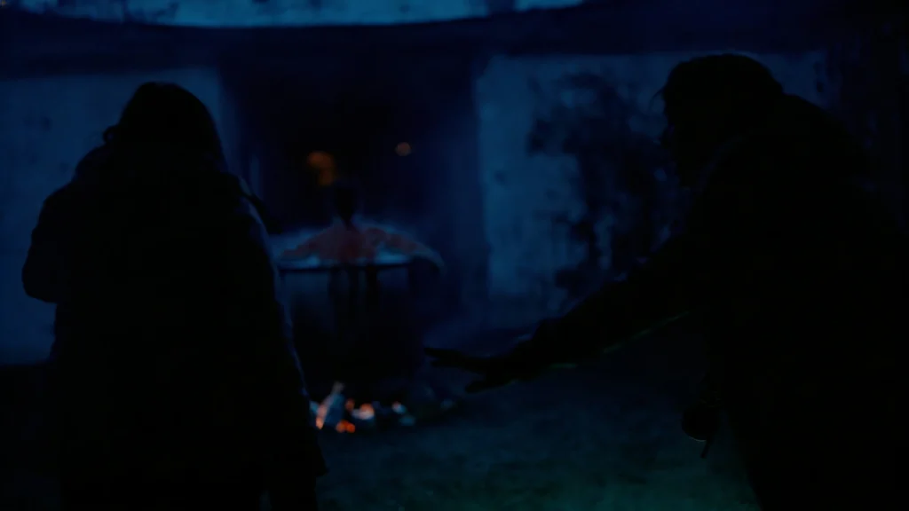 Two silhouetted figures face a smoking cauldron and a seated man in a blue-lit concrete interior.