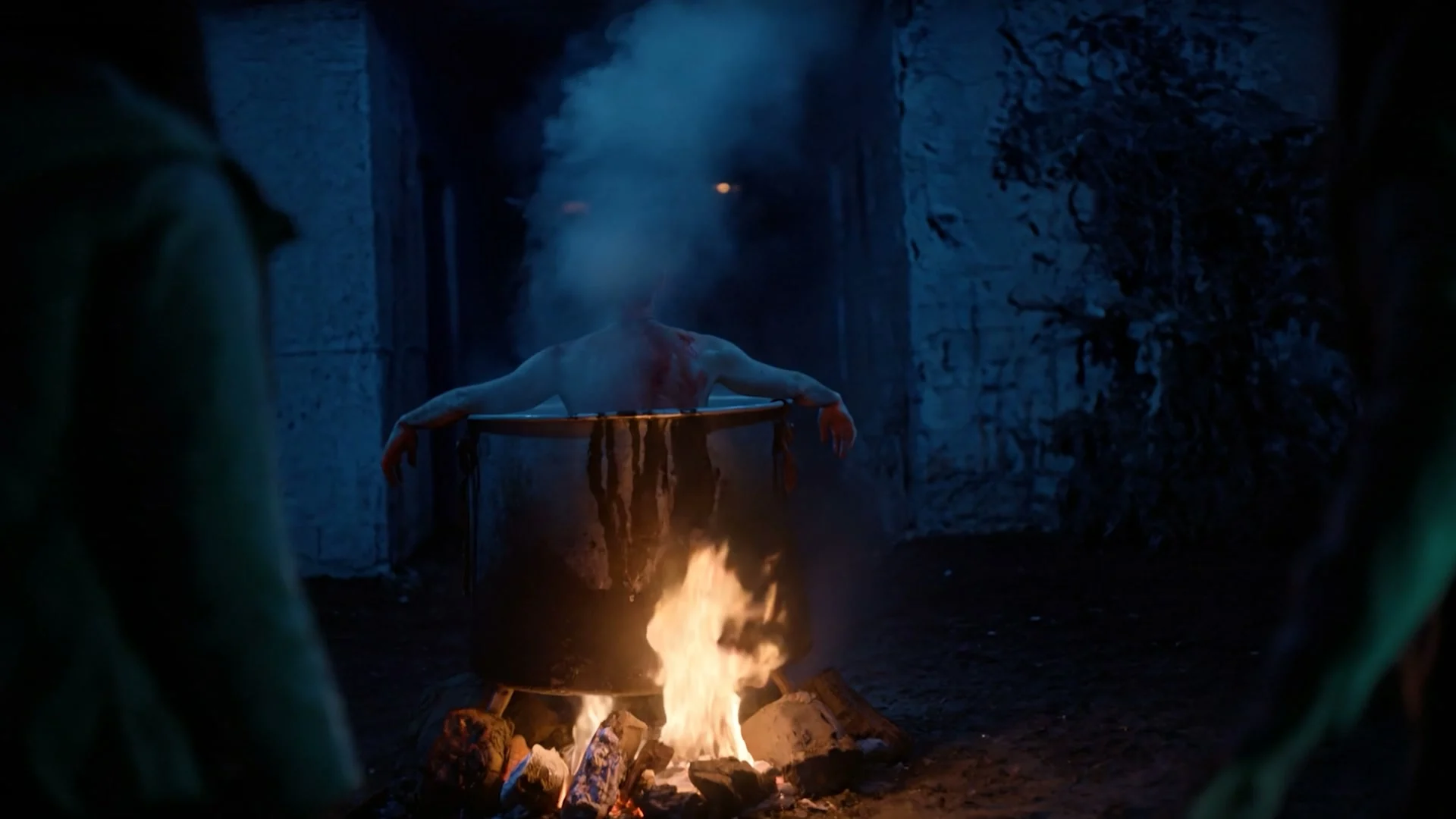 A man sits in a smoking cauldron above orange flames, framed by shadowed figures.