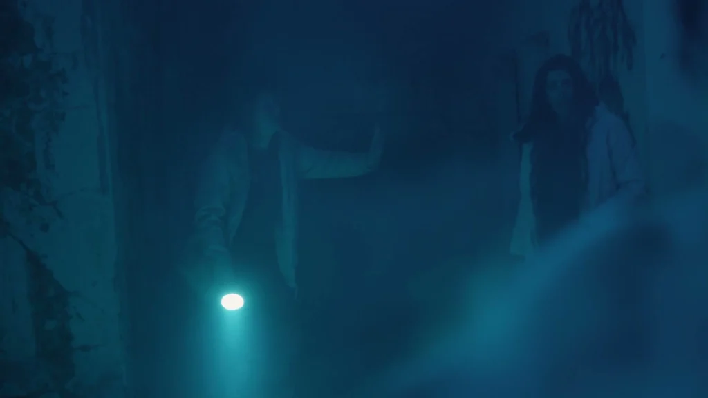 A man holds a flashlight beside a woman in a smoke-filled, blue-lit concrete interior.