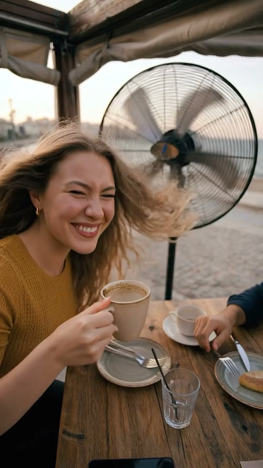 A laughing woman with coffee at an outdoor table, her hair blown by a Vanti pedestal fan: a frame from a Vanti social media commercial